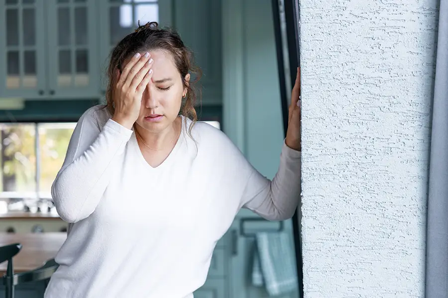 Woman leaning against a wall with her hand to her forehead and eyes closed, experiencing a headache or dizziness at home.
